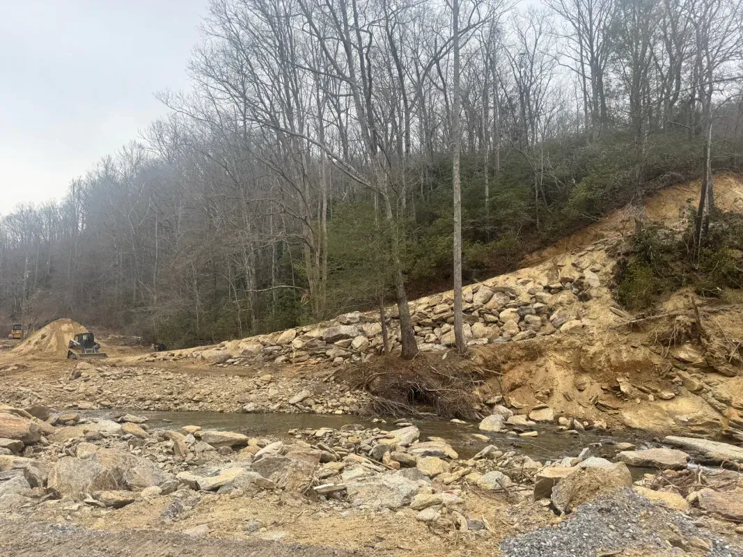 A landscape showing a muddy, excavated creek bed, a rocky slope, and a piece of construction equipment near a forest.