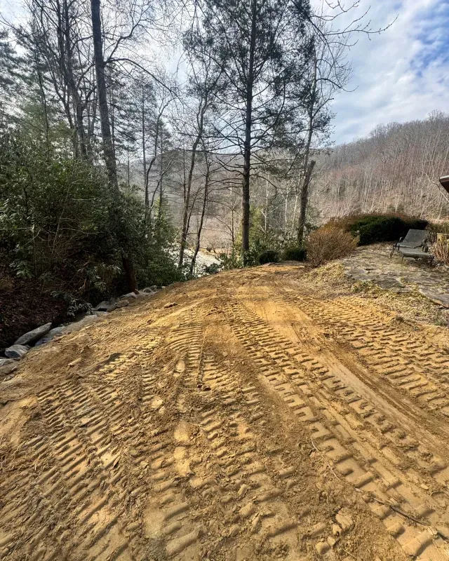 Freshly graded dirt slope with deep excavator tracks leading toward a wooded valley and hillside under a cloudy sky.