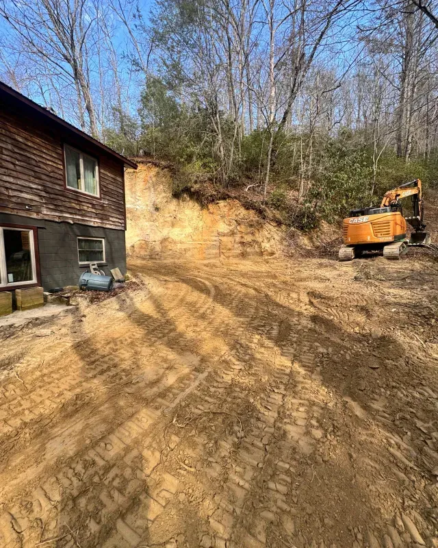 A yellow excavator parked on cleared dirt next to a wooden house with a steep, freshly excavated hillside behind it.