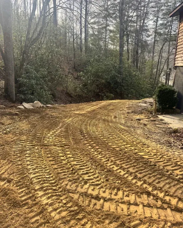Freshly graded dirt driveway with heavy machinery tire tracks, leading toward a wooded area near a house exterior.