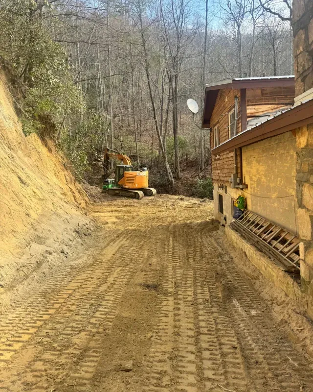 Yellow excavator parked on a dirt path between a steep, excavated earthen slope and a rustic wooden house.
