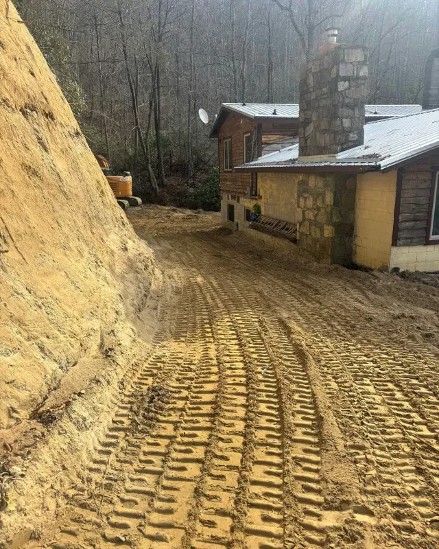 A dirt driveway with heavy machinery tire tracks leads toward a rustic cabin with a stone chimney in a wooded area.