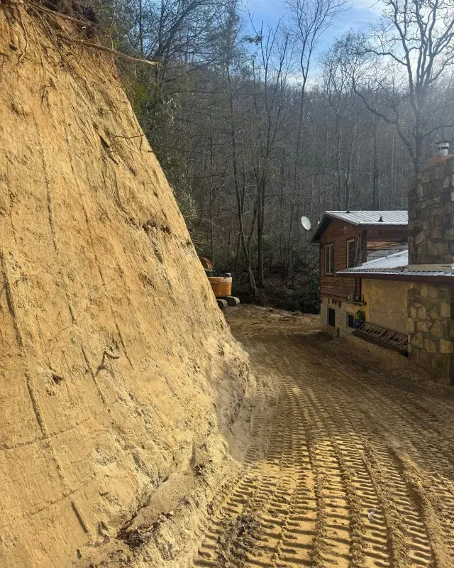 A steep, excavated earthen embankment beside a house with heavy machinery tracks in the dirt driveway.