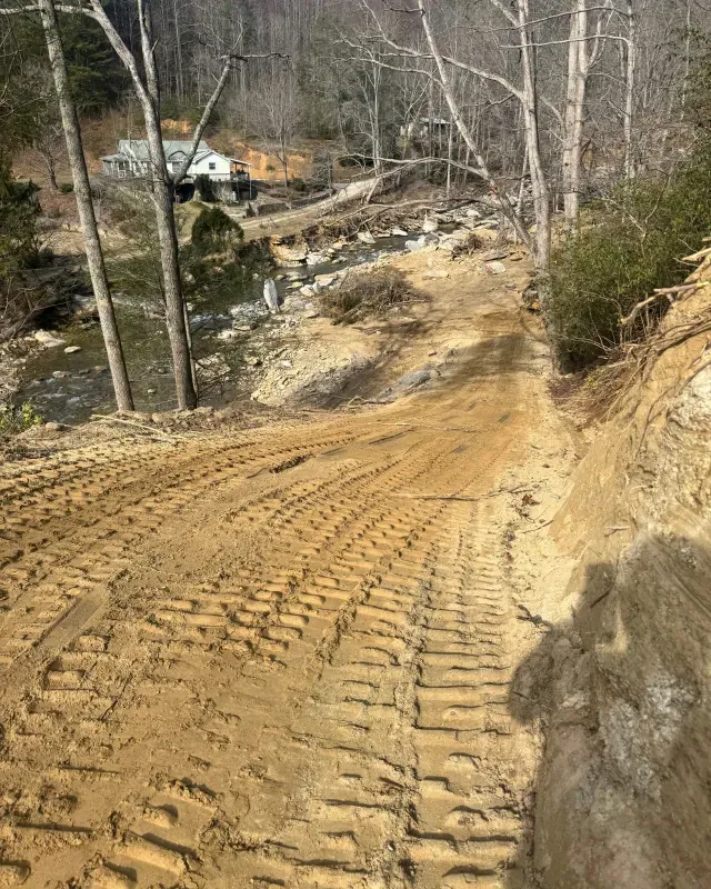 A steep, dirt slope with deep excavator track marks leads down toward a rocky stream near a house in a wooded area.