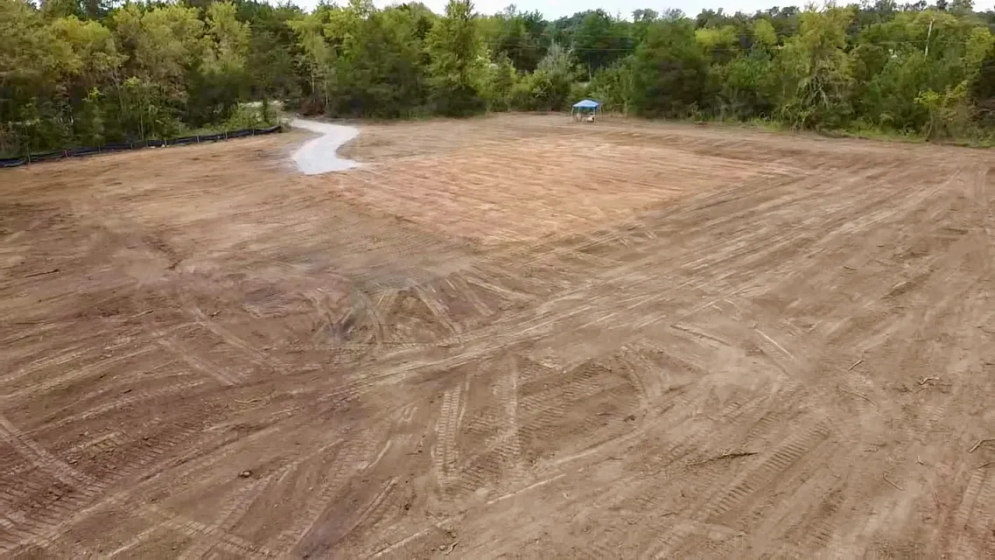 An aerial view of a cleared dirt lot surrounded by trees, showing tire tracks and a small gravel path in the corner.