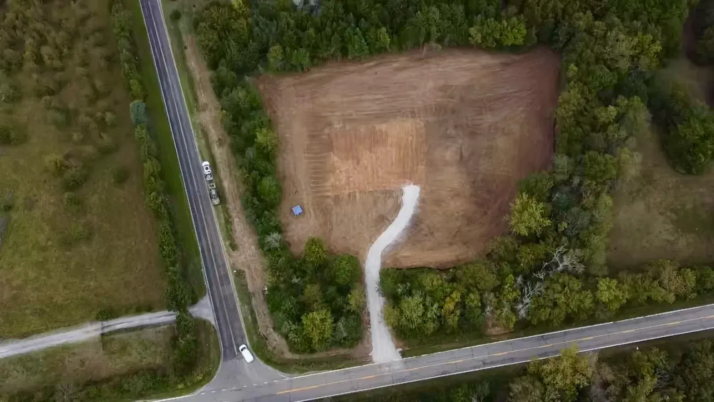 An aerial view shows a cleared, brown rectangular lot with a gravel driveway, surrounded by trees and two rural roads.