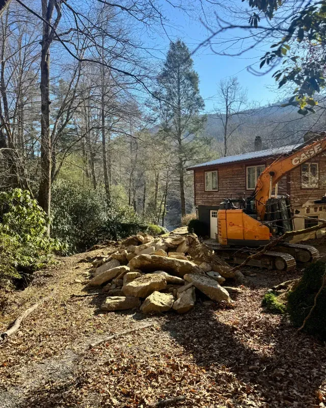 An orange excavator sits next to a pile of rocks in front of a wooden house nestled in a wooded, mountainous area.