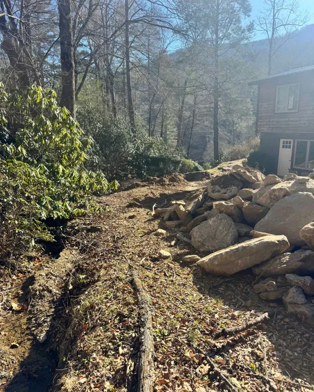 A dirt path winds through a forest beside a large pile of rocks and a portion of a wooden house under a bright blue sky.