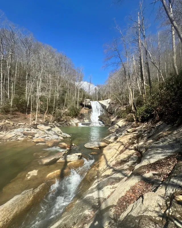 A waterfall cascades into a calm pool surrounded by bare, leafless trees under a clear blue sky.