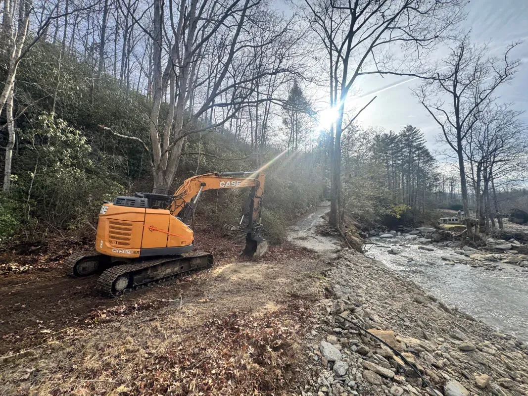 An orange excavator works on a dirt bank beside a flowing river in a wooded area during the day.