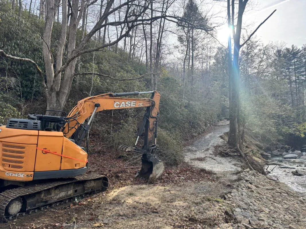 An orange Case excavator sits on a dirt path next to a stream in a wooded area.