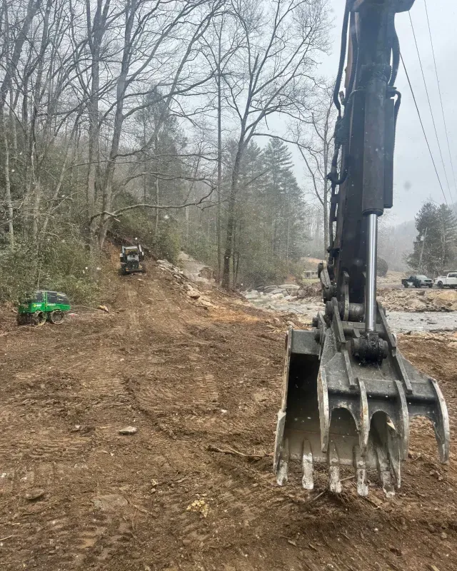 A close-up of a large black excavator claw with a construction site, trees, and heavy machinery in the background.
