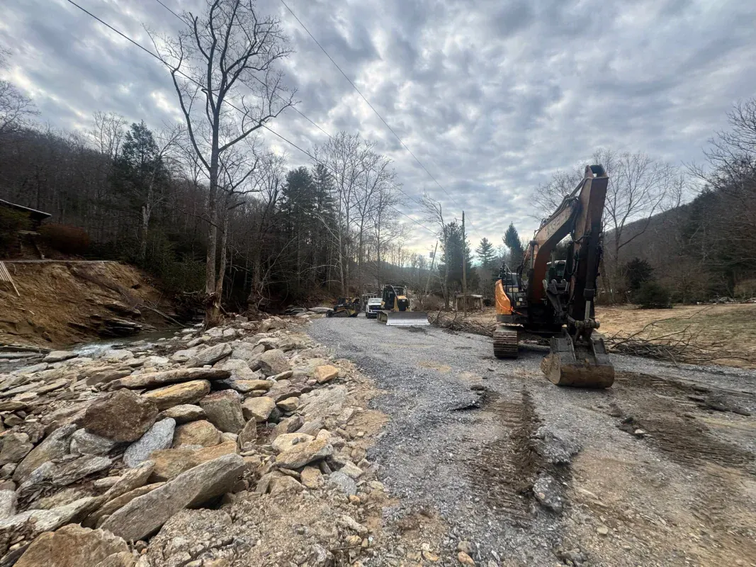 An excavator clears a debris-strewn road beside a forest stream under a cloudy sky.