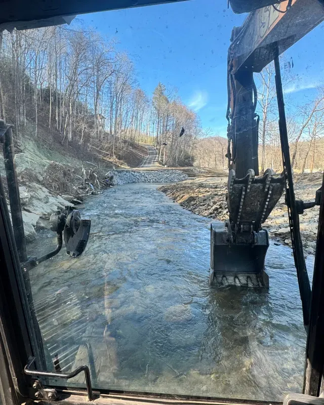 A view from an excavator cab showing the heavy machinery standing in a shallow, clear stream near a wooded bank.
