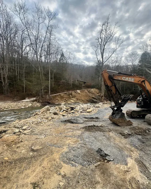 A yellow Case excavator parked on a dirt bank next to a river with a wooded, cloudy background.
