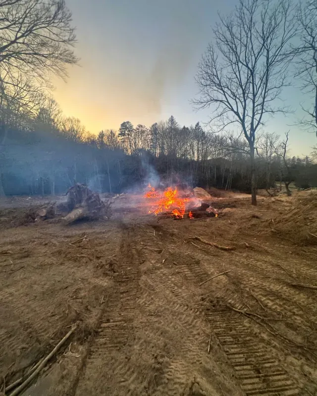 A brush pile burns in a cleared dirt field at sunset, with a line of bare trees in the background.