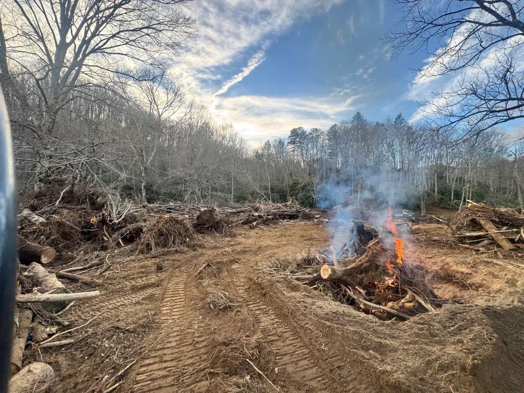 A bonfire burns amidst a landscape cleared of trees, with mud tire tracks visible in the dirt foreground.
