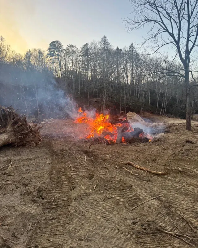 A bonfire burns in a cleared dirt lot near a forest at dusk, with smoke drifting into the sky.