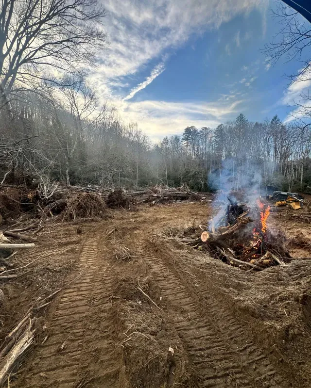 A bonfire burns on a cleared, muddy lot with heavy equipment tracks in the dirt, set against a backdrop of bare trees.