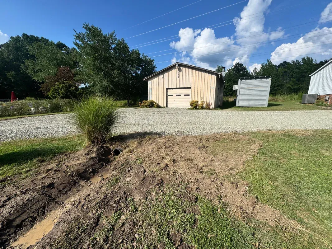 A wooden garage and a white shed sit beyond a gravel driveway, with a muddy trench in the foreground.