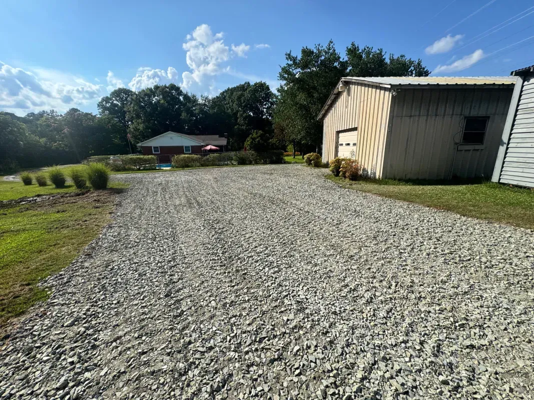 A gravel driveway leads toward a light-colored barn and a small house set against a treeline under a bright blue sky.