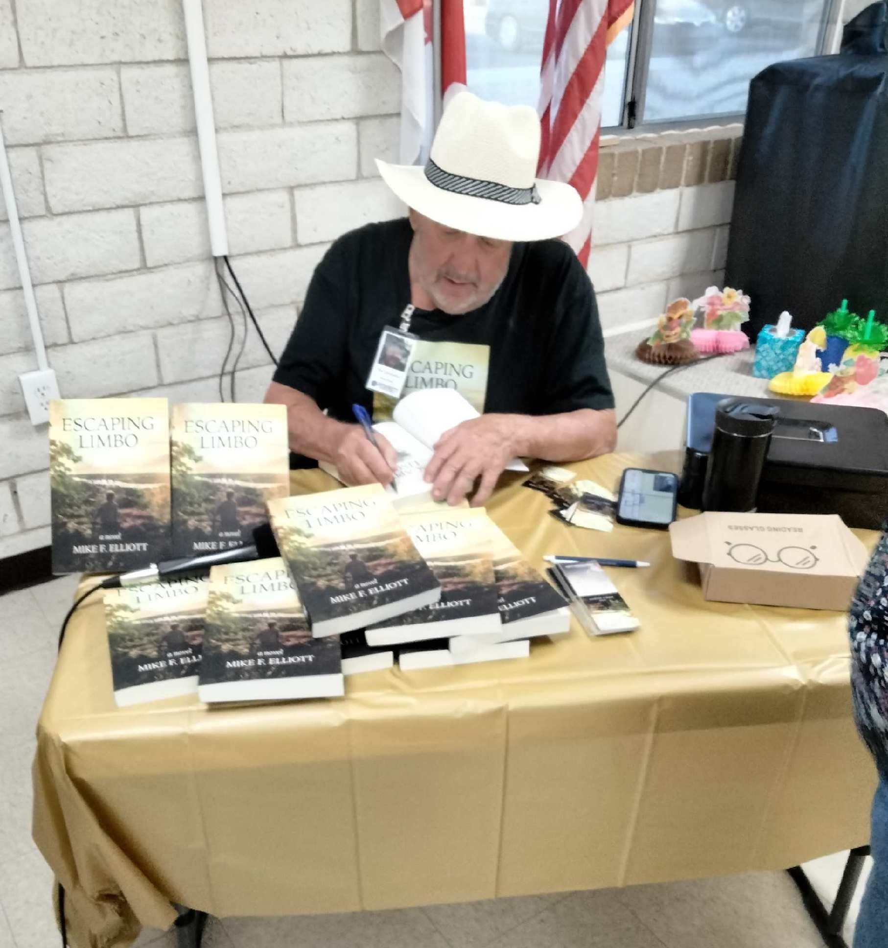 Author signing books at a table. Books, pen, phone, and food container are present.