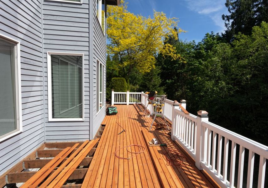 Wooden deck under construction next to a house with gray siding. White railing and green trees in the background.