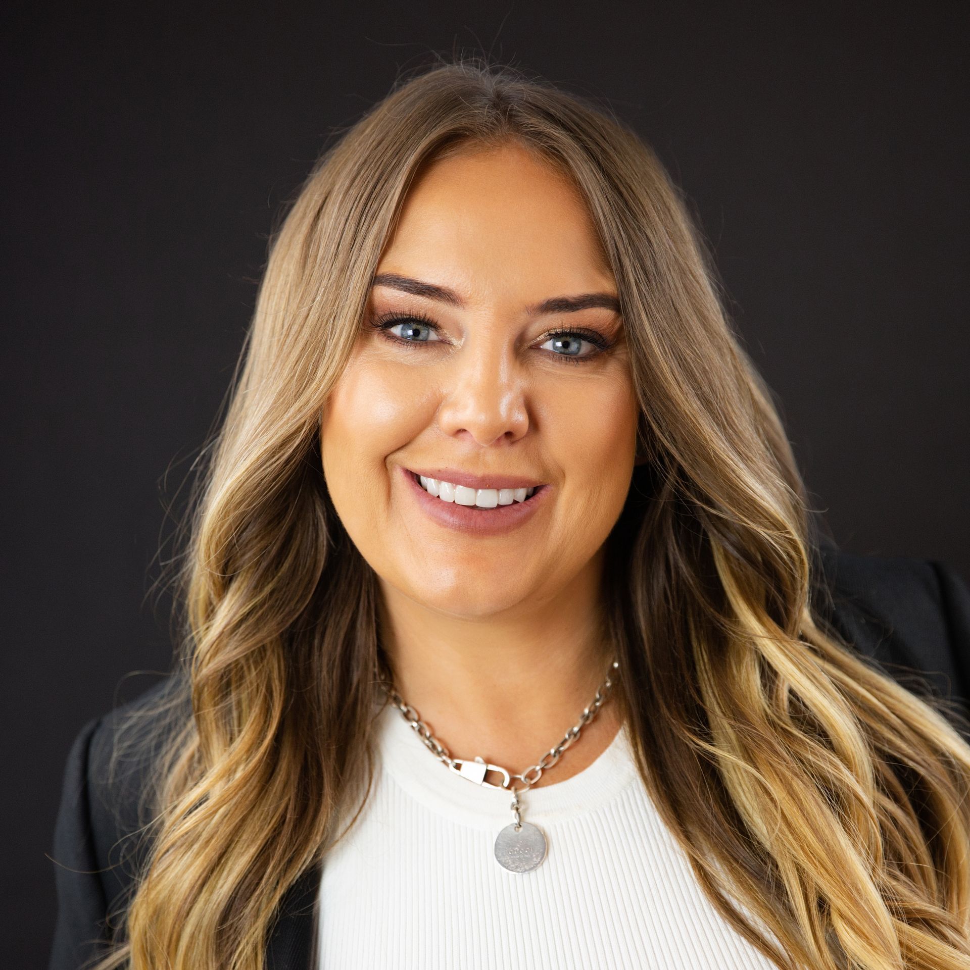 Woman with long blonde hair, smiling, wearing a white sweater, black blazer, and silver necklace against a black background.