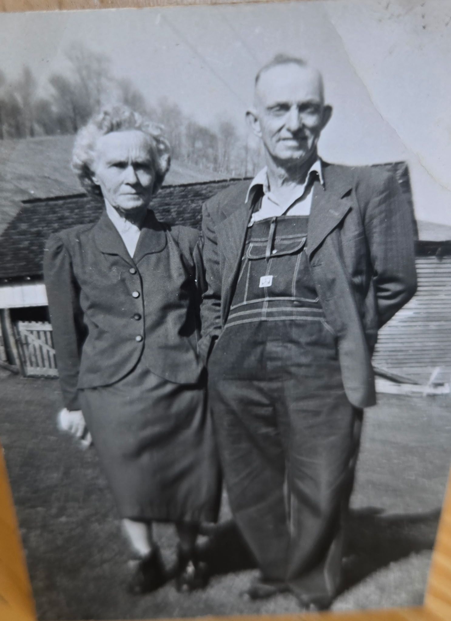 Elderly couple standing outside, the man wearing overalls, the woman in a suit.