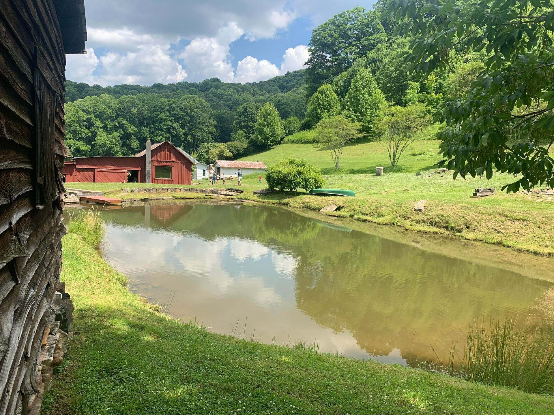 A pond reflects the sky in front of a red barn, white building, and green hillside on a sunny day.