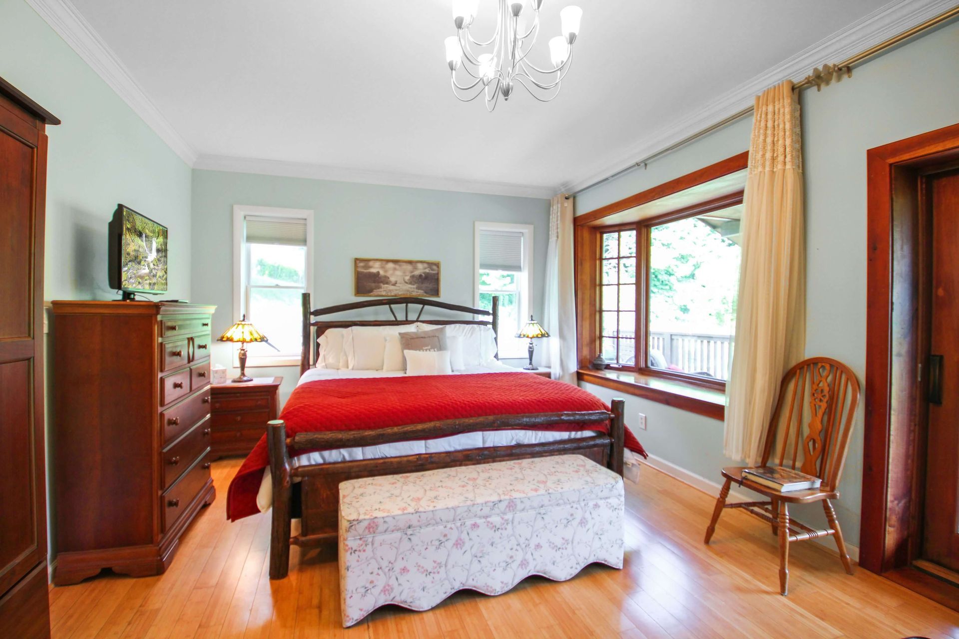 Bedroom with bed, chest of drawers, and bay window. Red bedspread, hardwood floors, light blue walls, and wooden furniture.