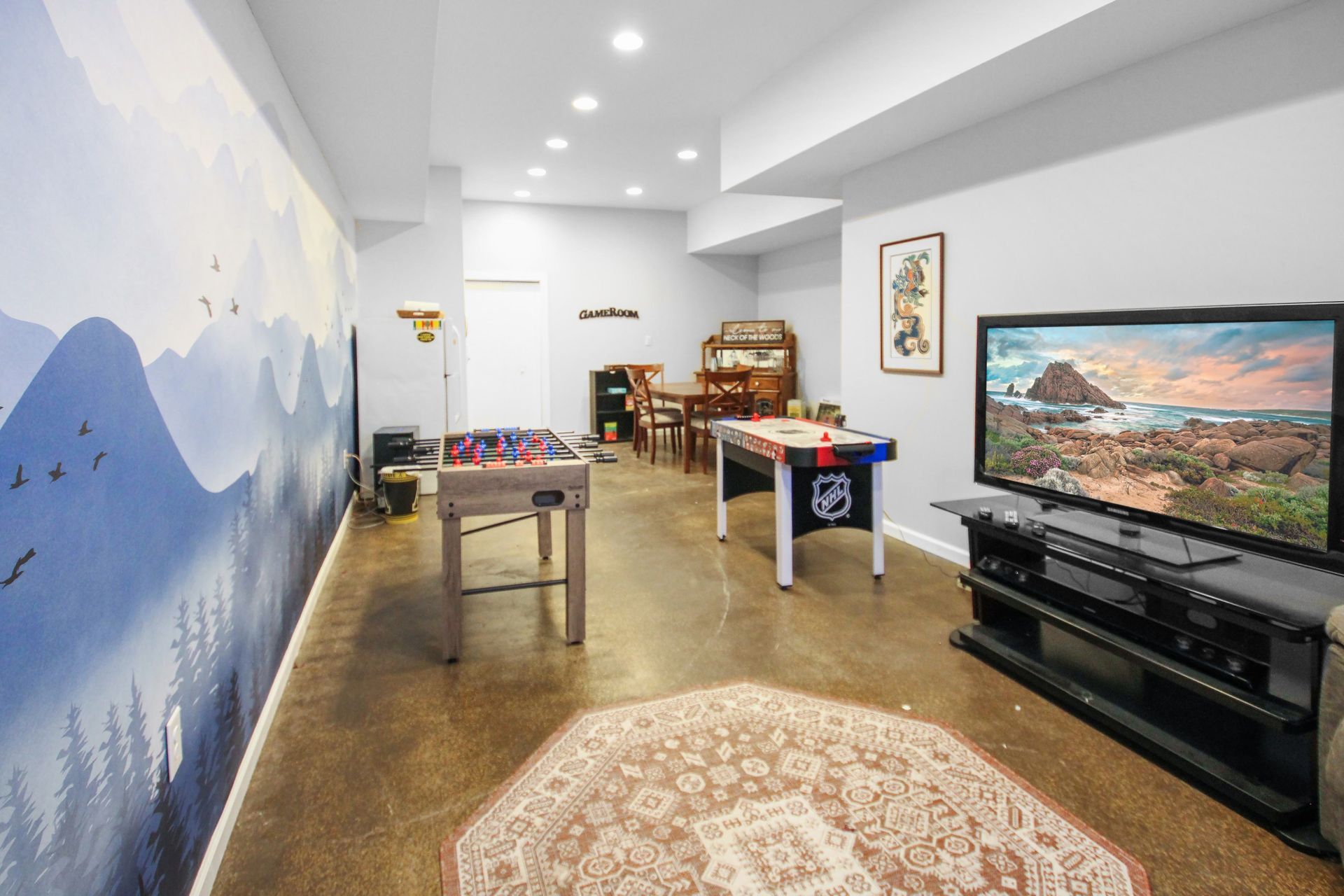 Basement with foosball, air hockey, and TV. Mural on wall, rug on floor, and a dining table.