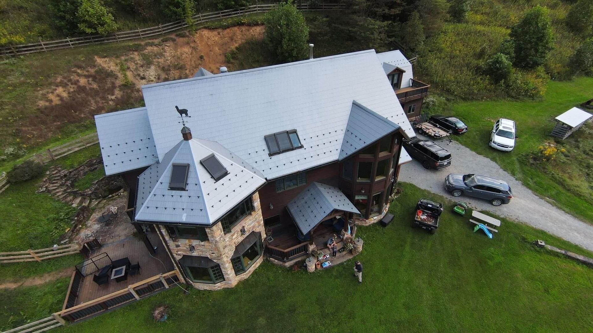 Drone shot of a large house with a silver roof, surrounded by grass and parked cars.