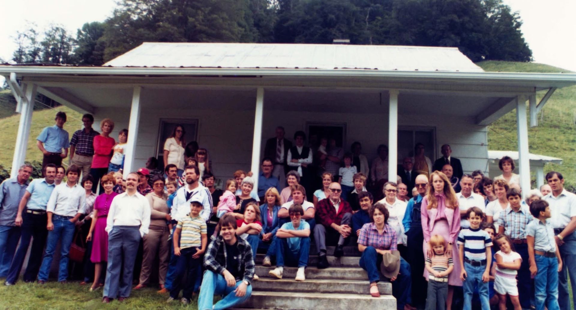 Large group of people posing in front of a white house with a porch. Outdoors on a hill.
