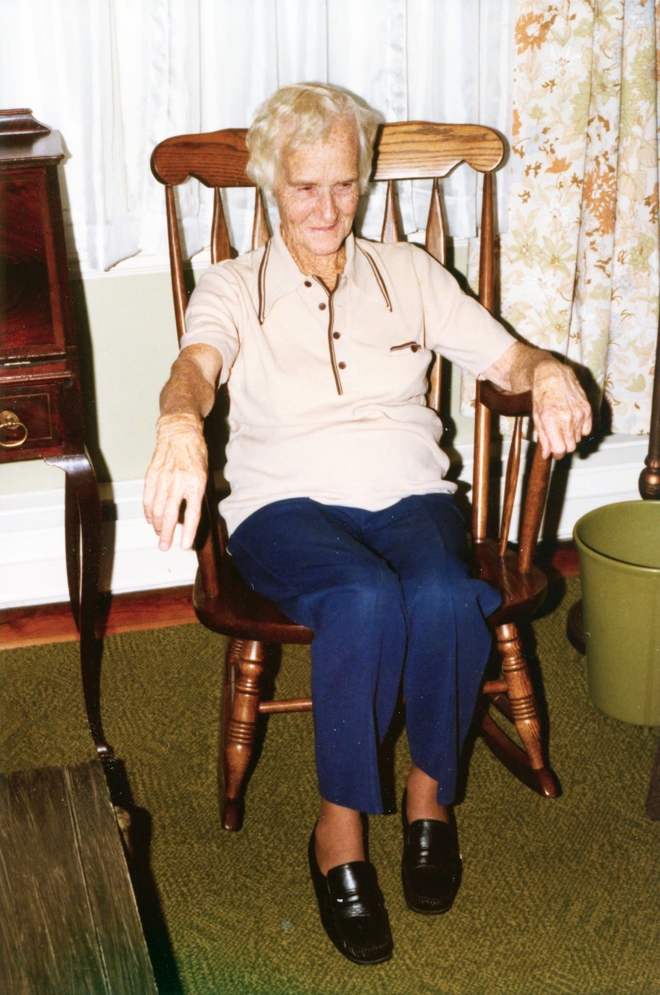 Elderly woman sitting in a wooden chair. She wears a light shirt, blue pants, and black shoes. Inside a room.