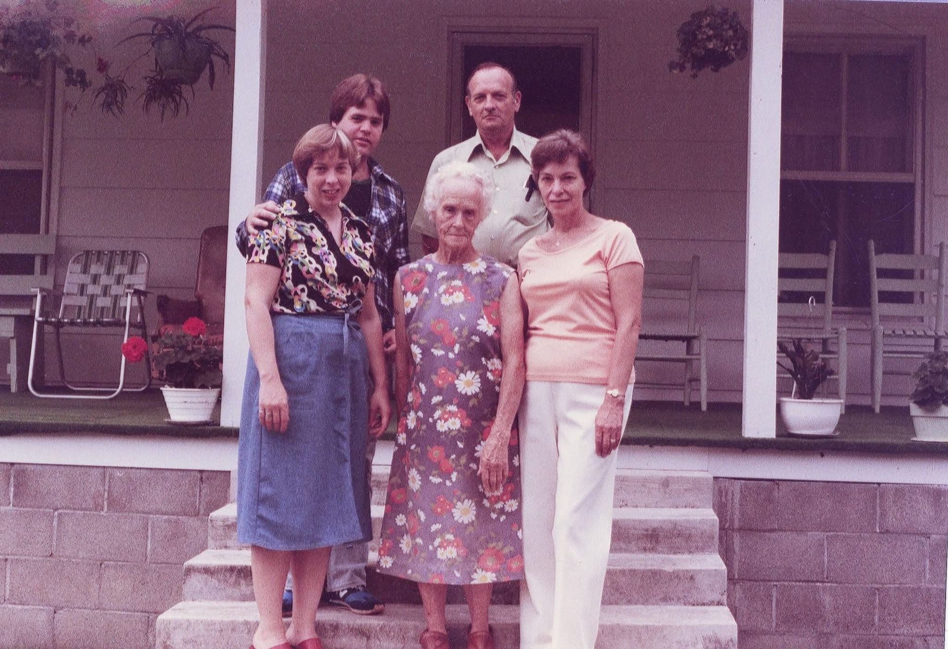 Family of five stands on porch steps; woman in floral dress at center.