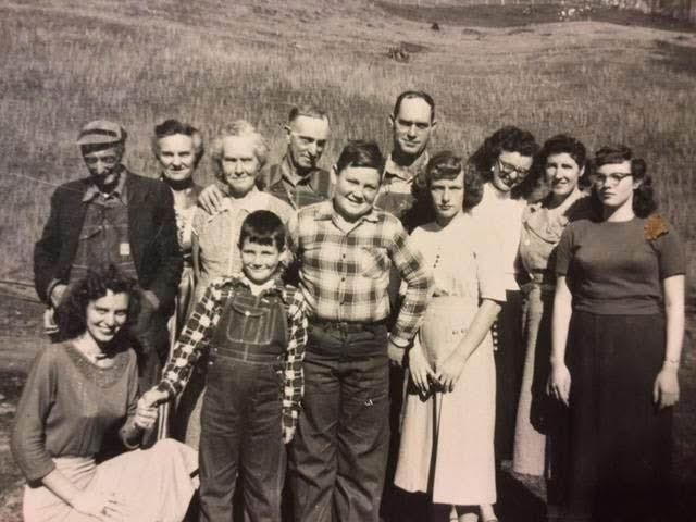 Group photo of a family posed outdoors. People smiling, wearing casual clothing. Overcast day, tall grass in background.