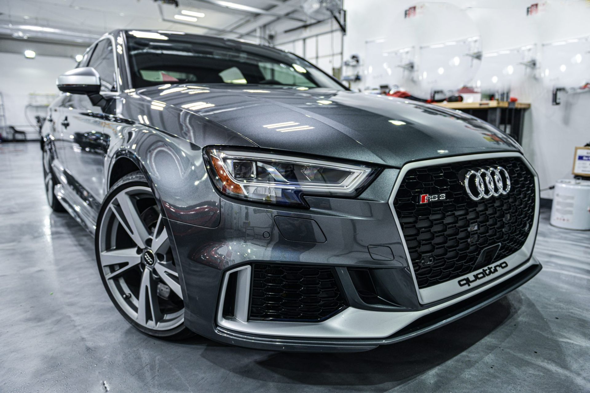 Gray Audi RS3 sedan in a bright garage, front three-quarter view with black grille and large alloy wheels