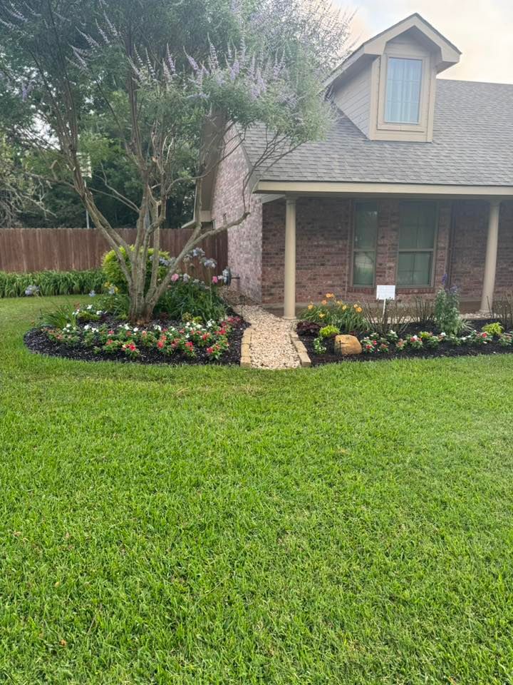 House with brick exterior, porch, and landscaped flower beds. Green lawn, trees, and walkway leading to the front door.