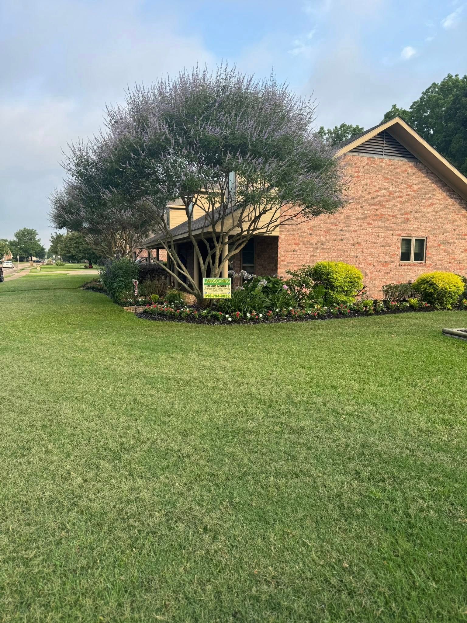 Brick building with a large tree in front, green grass, and a flower bed.