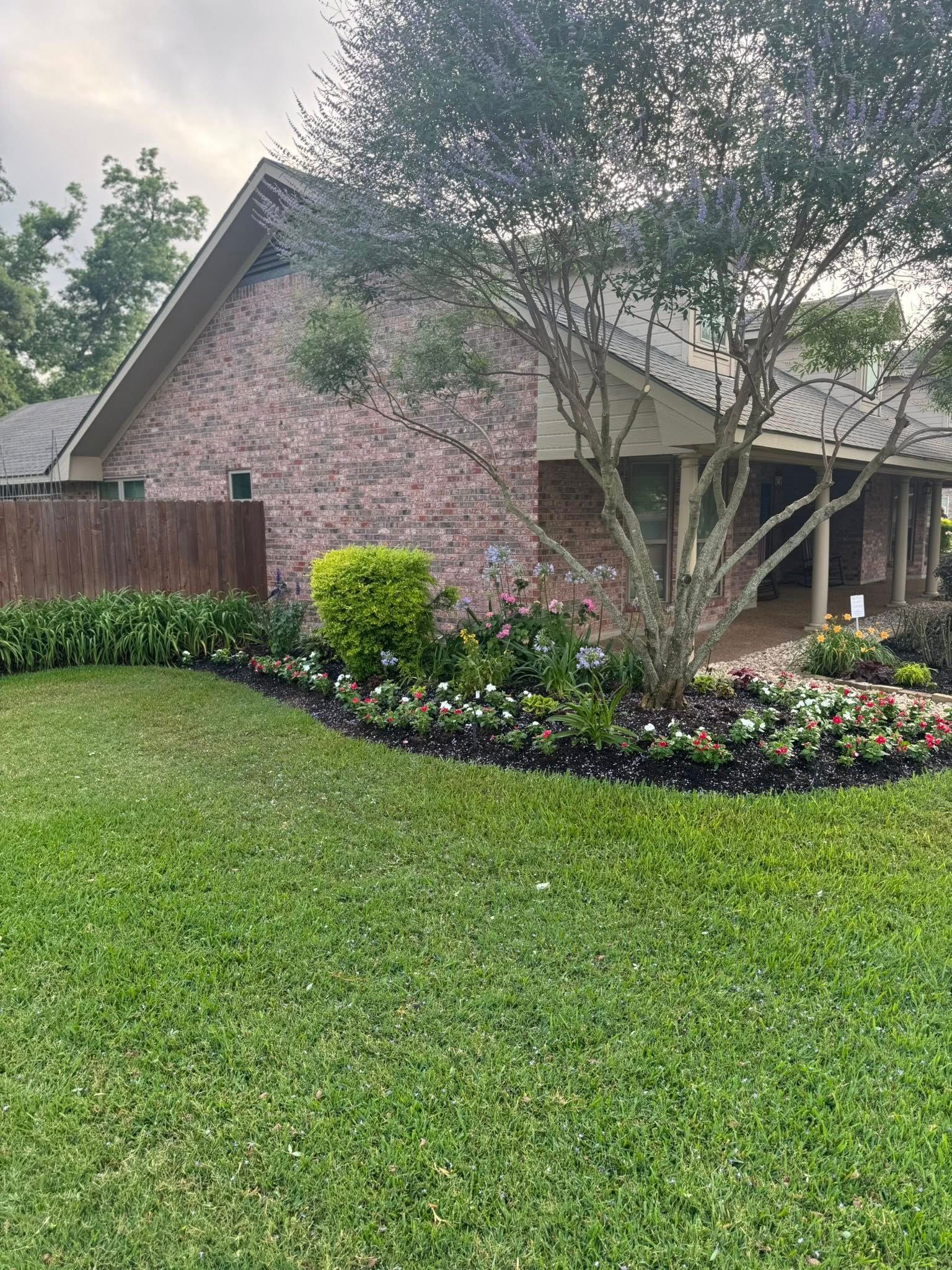 House with brick facade and landscaped flower beds. Green lawn, mature tree, and wooden fence.
