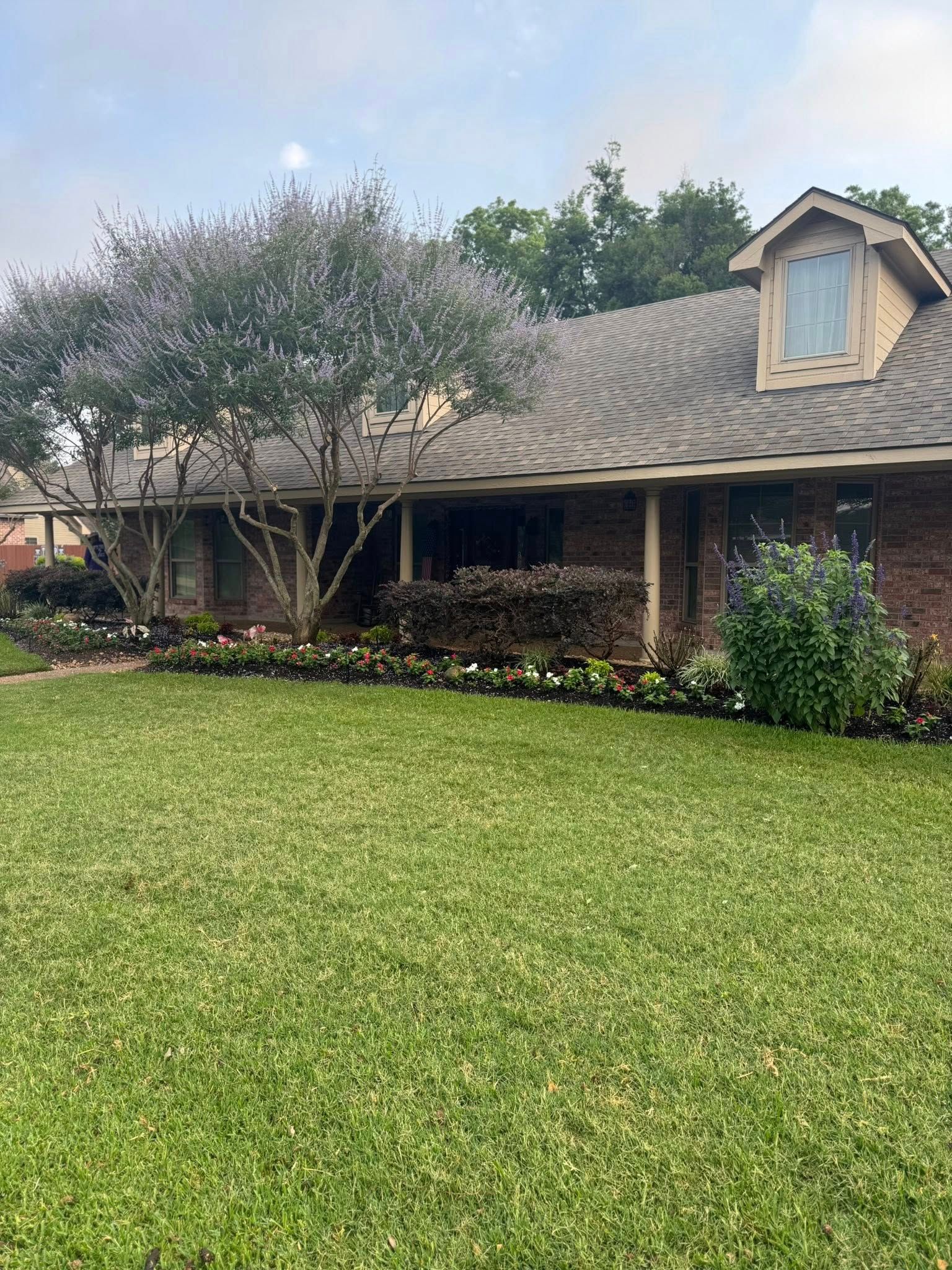 House with a manicured lawn and garden beds, mature trees, and a dormer.