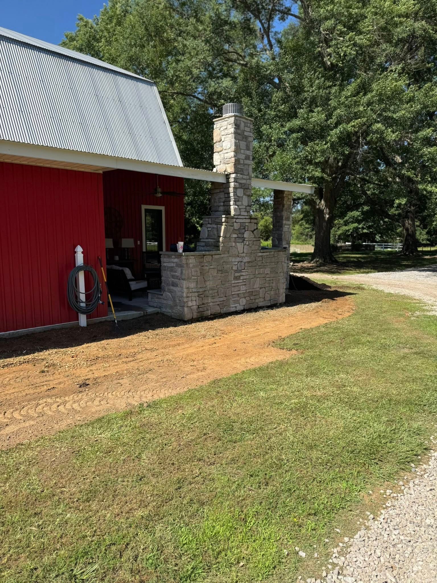 Red building with stone chimney and partial porch, next to gravel driveway and green grass.