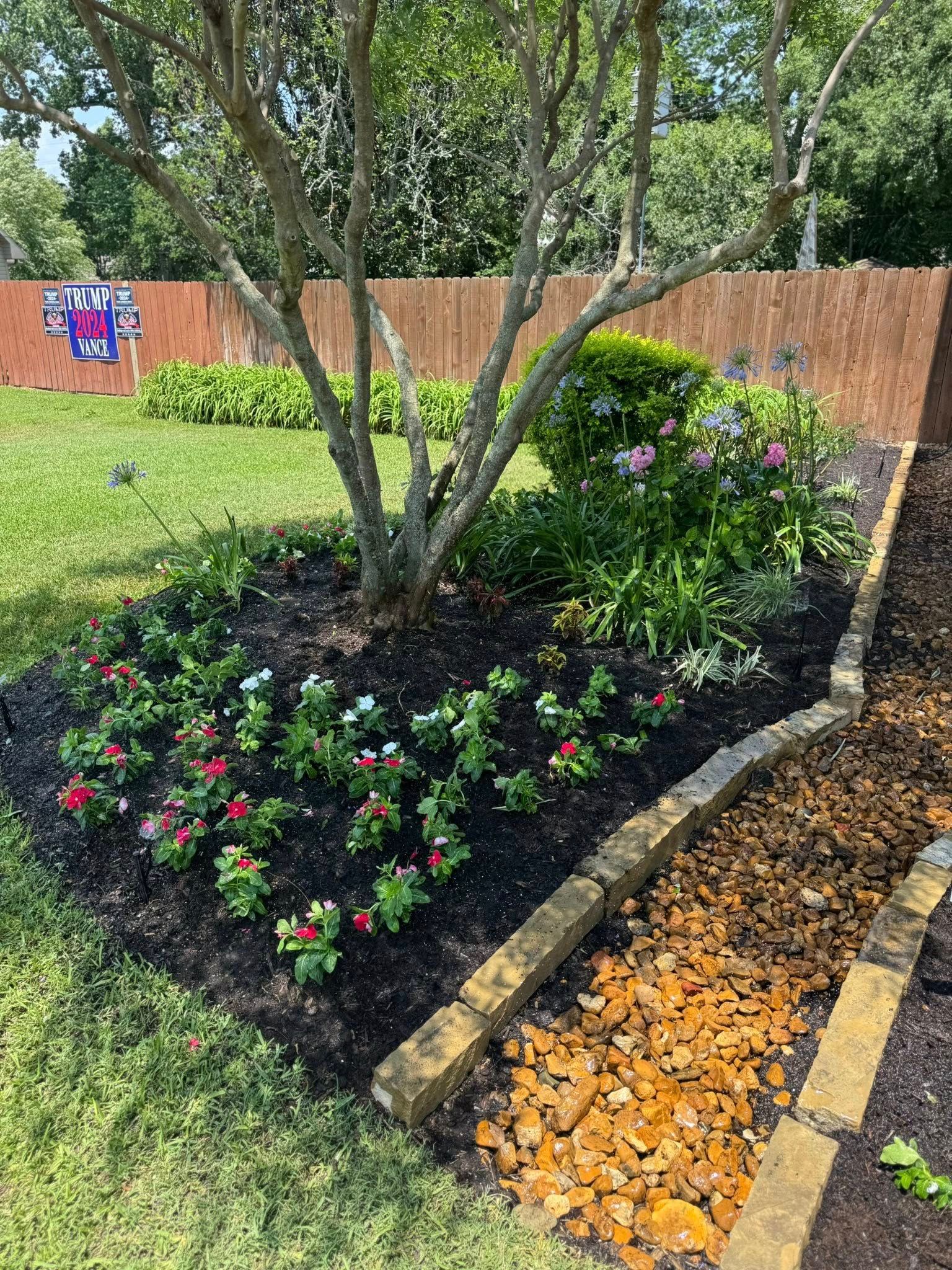 Flower bed around a tree with red, white, and pink flowers, bordered by bricks, with a wooden fence in the background.