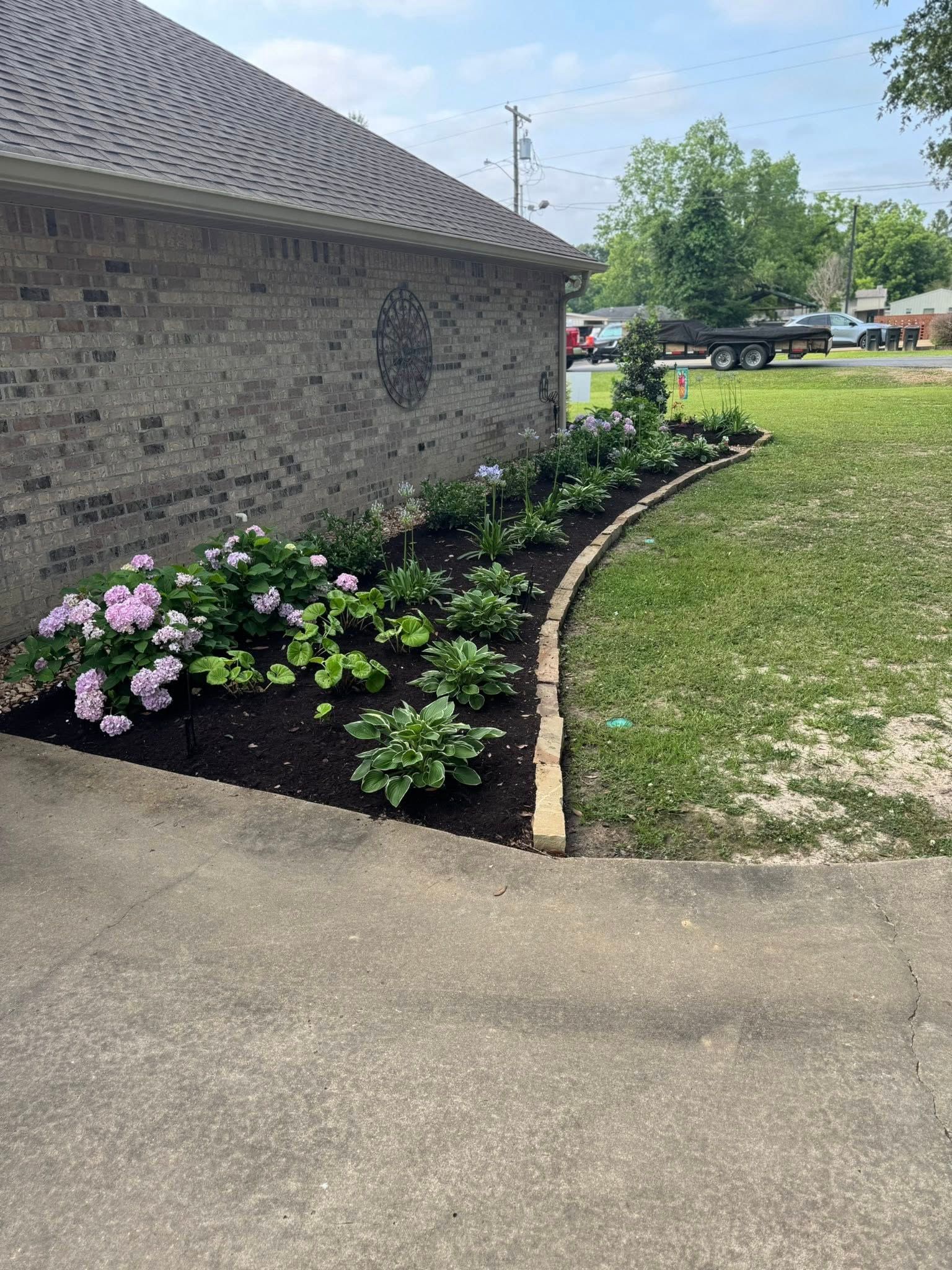 Brick building with a curved flower bed filled with plants and mulch, bordered by stones.