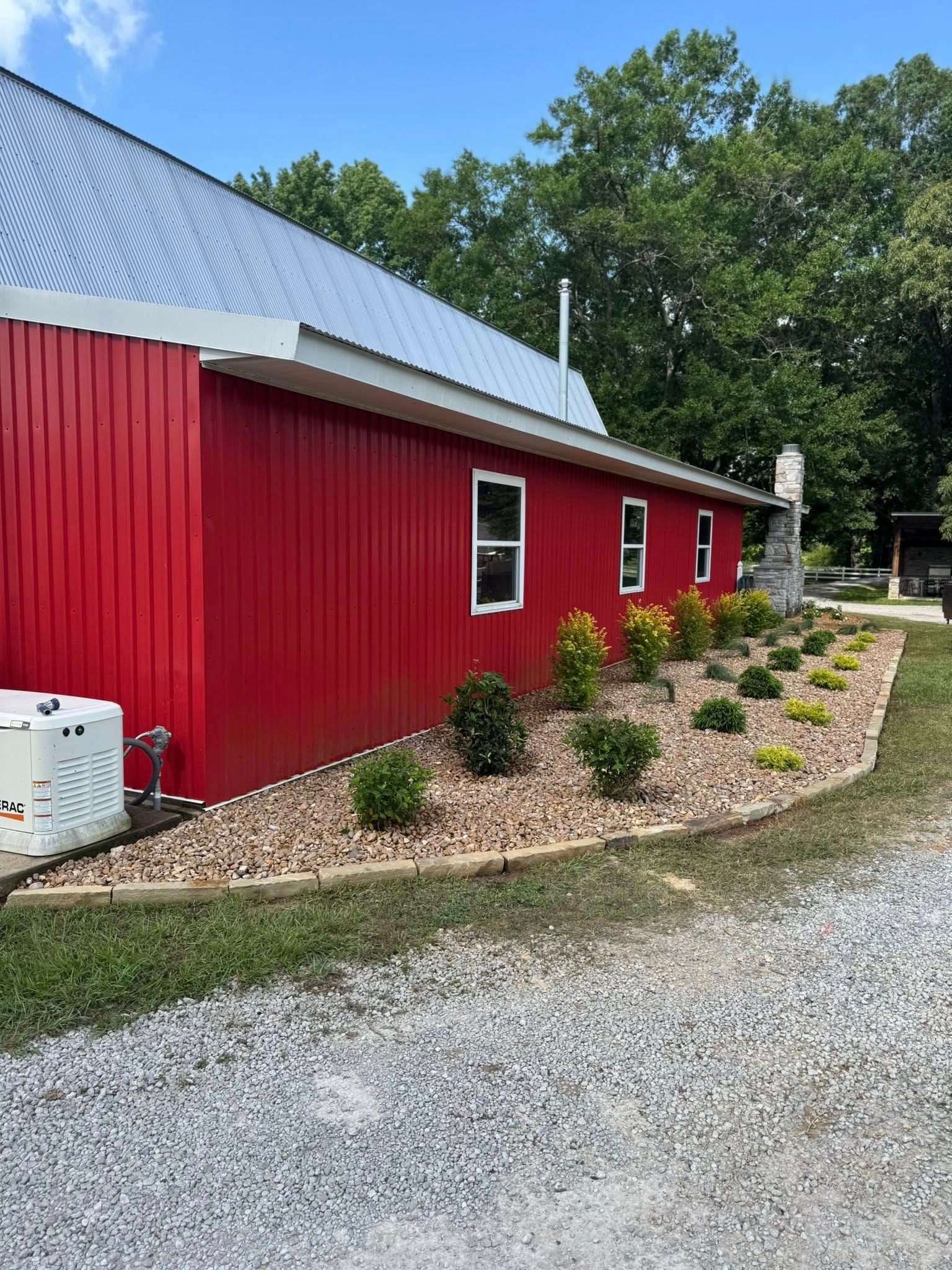 Red barn with a silver roof and small bushes in a gravel bed, with a gravel driveway.