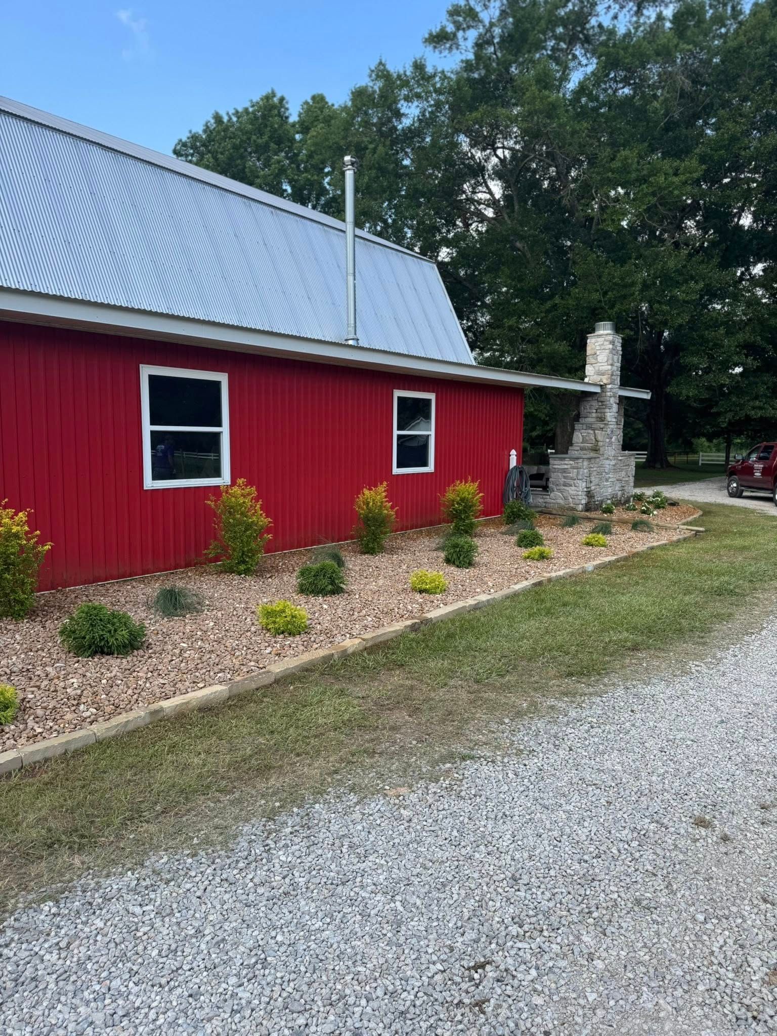 Red barn with a silver roof, white-framed windows, and a gravel driveway. Landscaped with small shrubs.