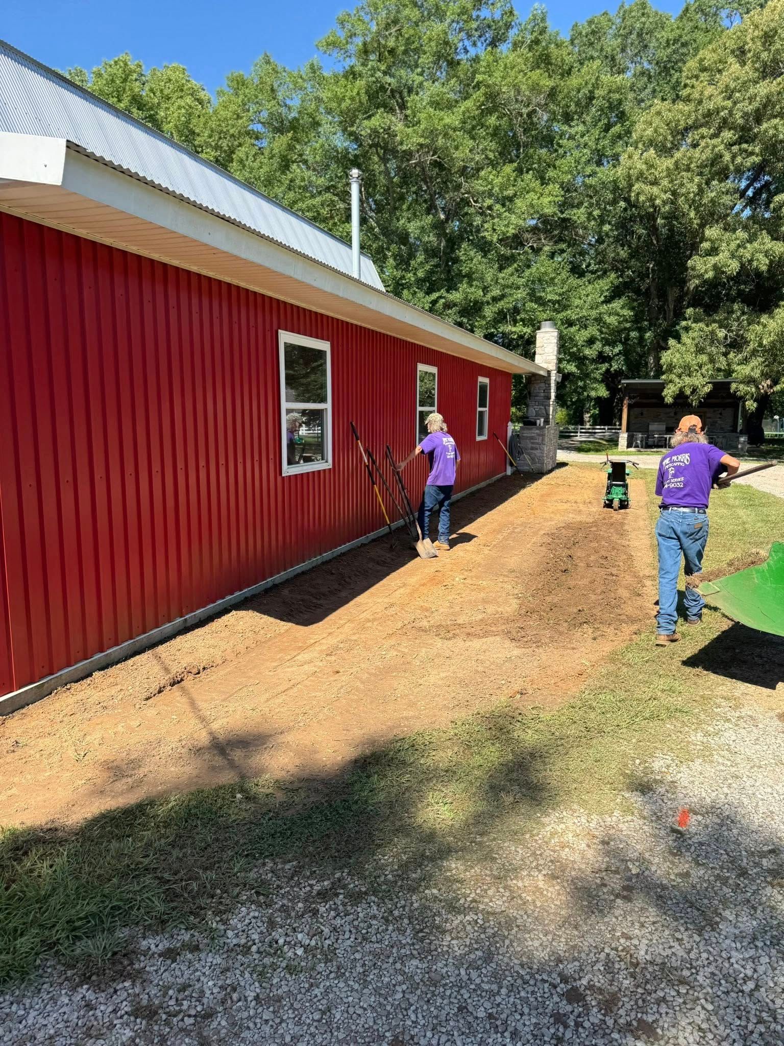 Two people in purple shirts work on a garden bed next to a red building with a metal roof.