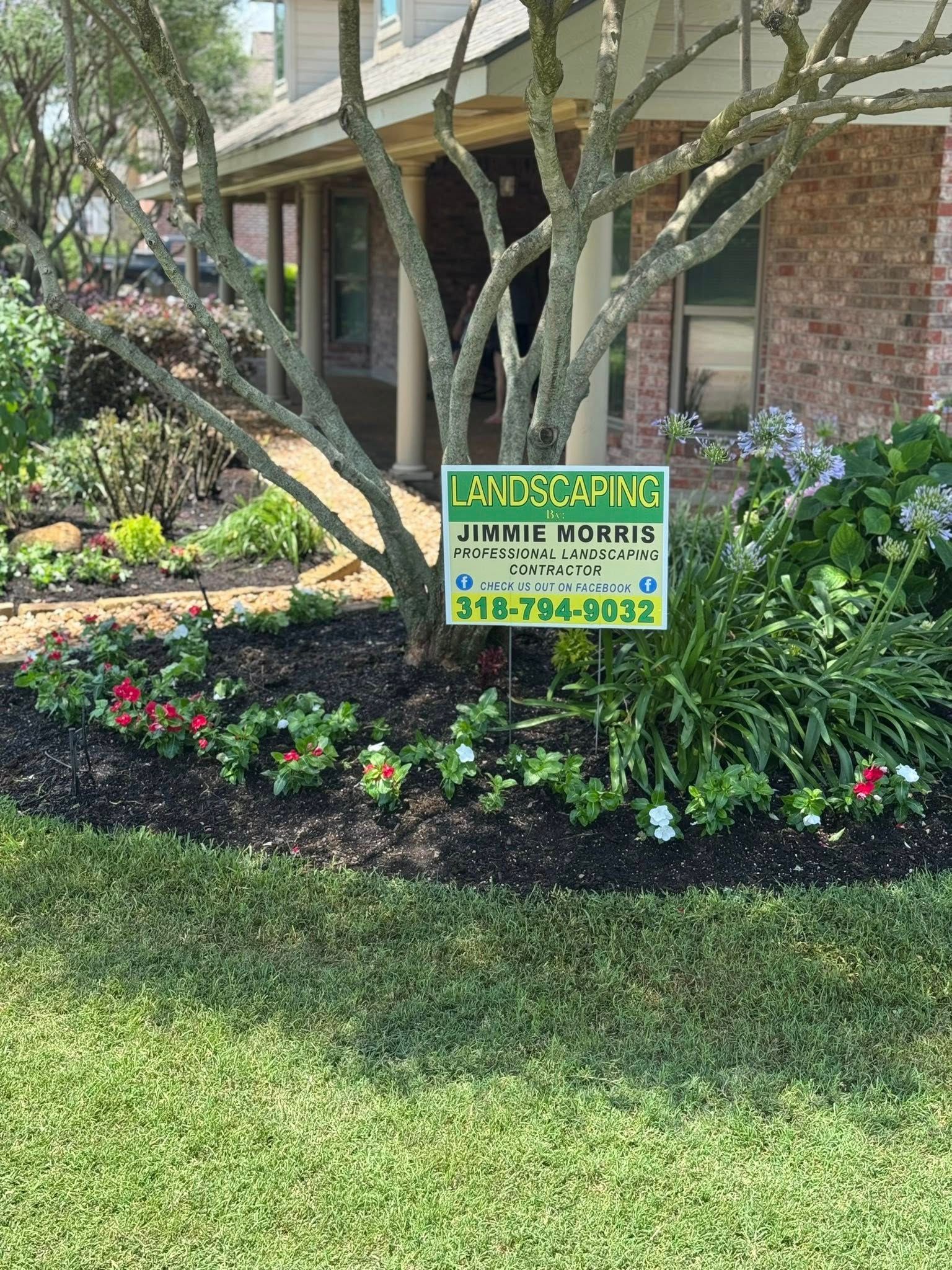Landscaping sign in a flower bed in front of a brick house. Green grass and flowers surround the bed.