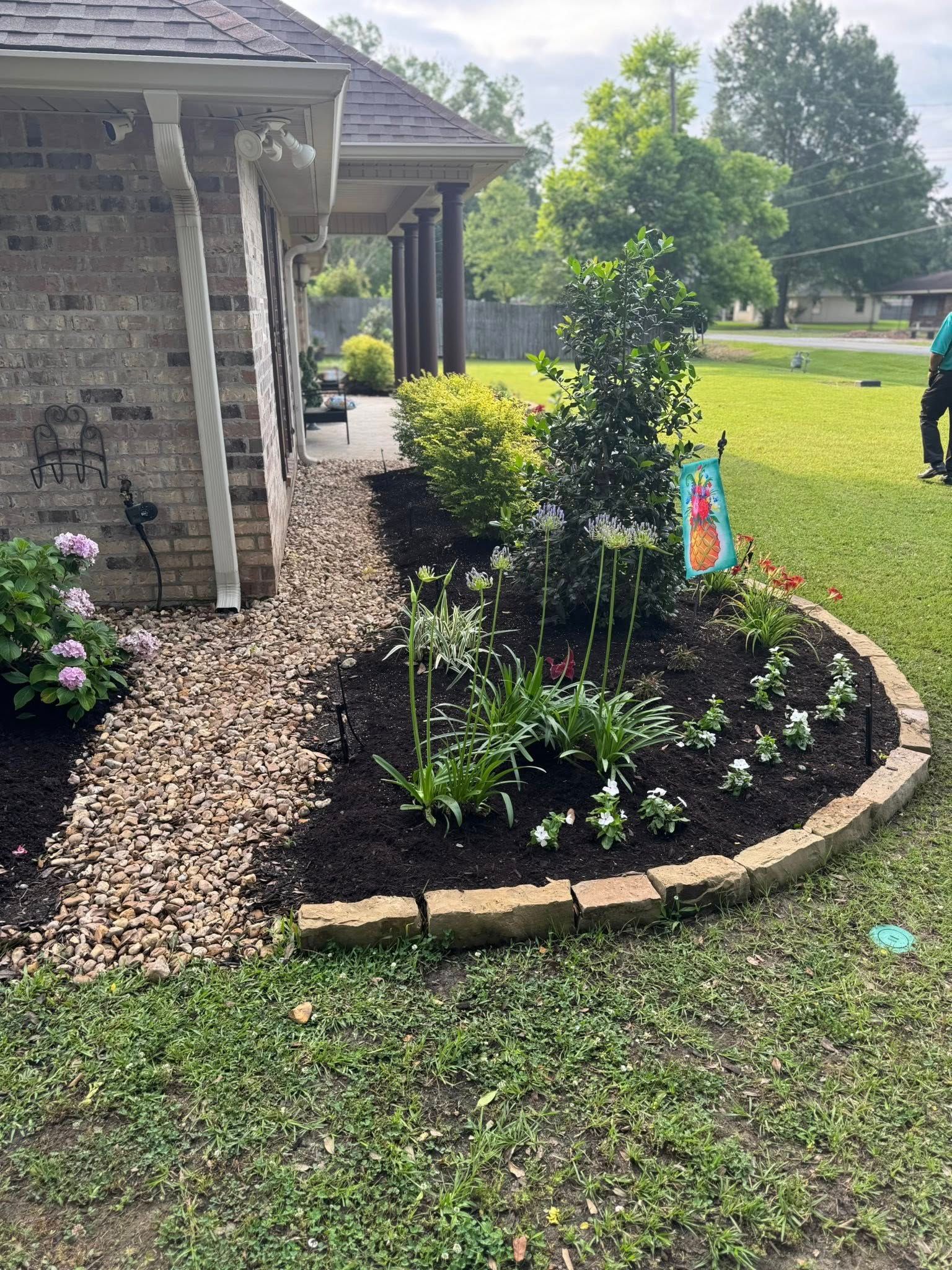 A flower bed with various plants and a decorative border in front of a brick house.